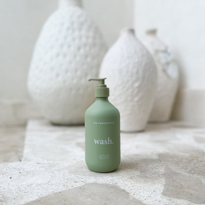 A bottle of body wash on a table beside a decorative vase filled with flowers.