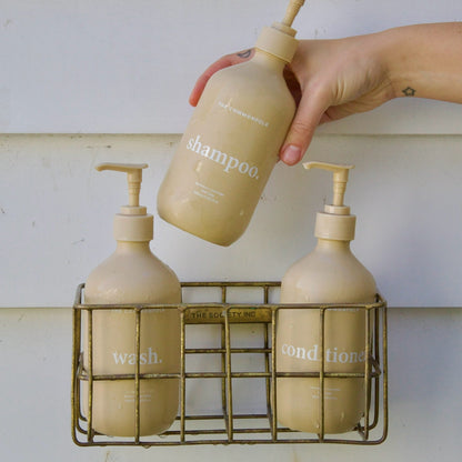 Hand holding a beige shampoo bottle over a wire caddy with other bottles on a white background