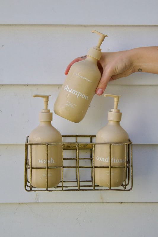 Hand holding a beige shampoo bottle over a wire caddy with other bottles on a white background