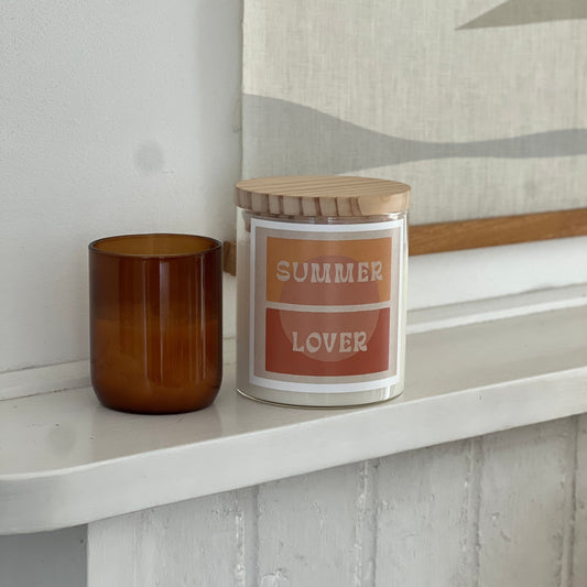 Candle labeled 'Summer Lover' on a shelf with a decorative painting in the background.