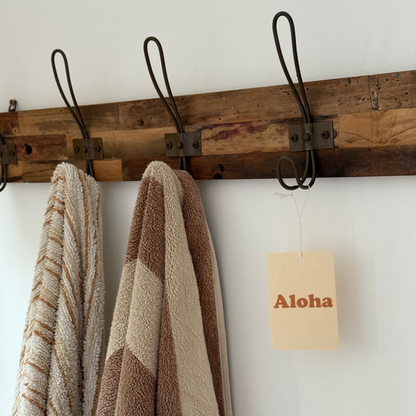 Wooden towel rack with towels and an air freshener 'Aloha' sign on a white wall.