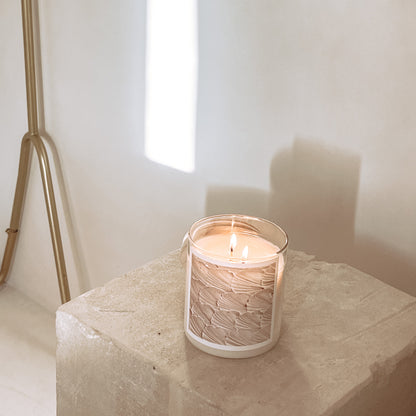 Candle in a textured glass holder on a stone surface with a white wall and decorative basket in the background.