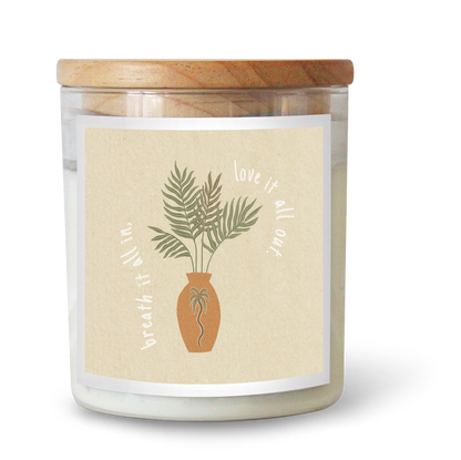 The Commonfolk scented white candle with a wooden lid, features a label with a palm plant design and the text 'breath it all in, love it all out'. The brand Roam Slow Studio is mentioned.