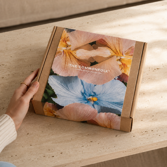 A hand holds a square cardboard gift box from The Combonfolk, decorated with a large floral print of peach and blue flowers, resting on a light wood table.