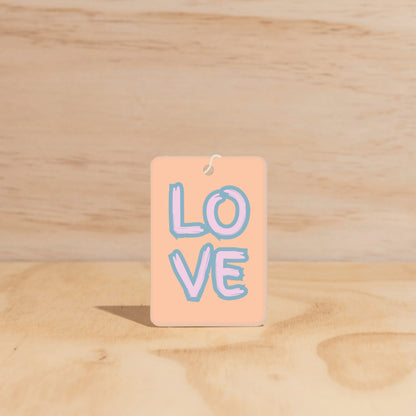 The Commonfolk air freshener with 'LOVE' written in blue on a beige background, hanging against a wooden backdrop.