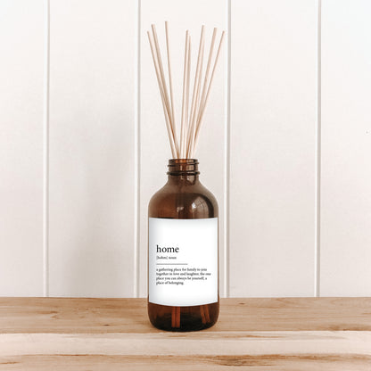 The Commonfolk room diffuser with an amber glass bottle and wooden reed sticks, labeled 'home', is placed on a wooden surface against a white background.