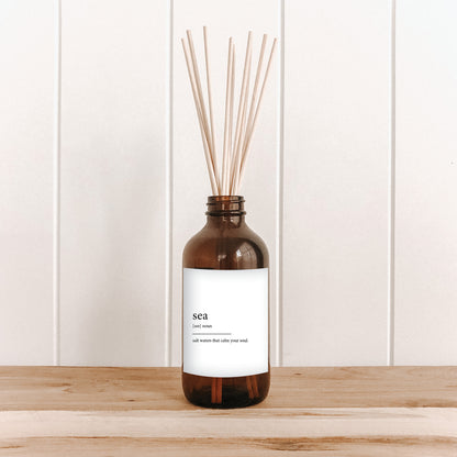 The Commonfolk room diffuser, with an amber glass bottle and bamboo reed sticks, and a white label, is placed on a wooden surface against a white background.