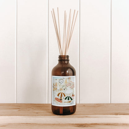 The Commonfolk room diffuser with natural bamboo reed sticks in an amber glass bottle features a label with an image of people on the beach.