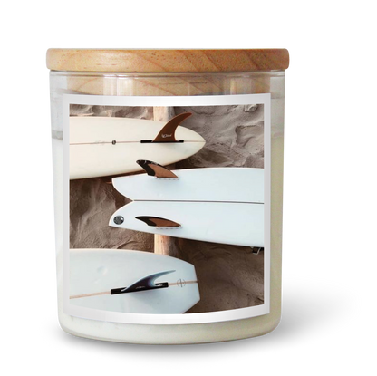 The Commonfolk scented candle has a white label inside a clear jar, featuring a wooden lid. The label shows a white surfboard.