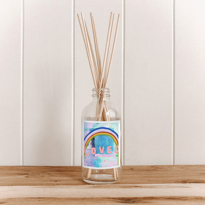 The Commonfolk room diffuser bottle, with a label featuring a rainbow and the word 'LOVED', filled with natural bamboo reed sticks, is placed on a wooden surface against a white background.