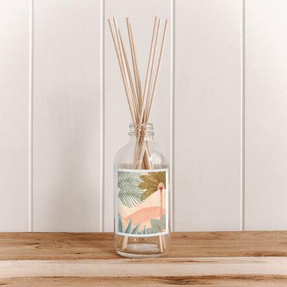 The Commonfolk room diffuser features a clear glass bottle with a colorful botanical illustration design, placed on a wooden surface against a white background.