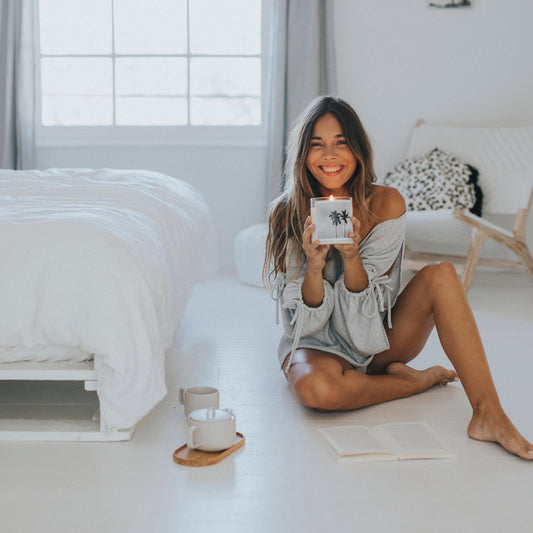 A woman sitting on the floor, holding a scented candle, appears relaxed and contemplative in a cozy setting.