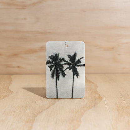 The Commonfolk air freshener, with a white background featuring black silhouettes of palm trees, displays on a wooden background.
