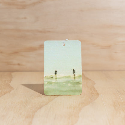 The Commonfolk air freshener, with a depiction of a beach scene, is displayed against a wooden background.