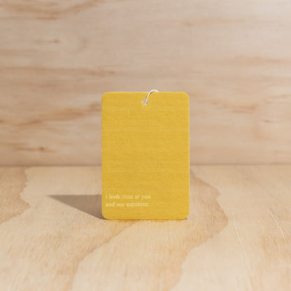 The Commonfolk air freshener features a yellow label with the text 'I look over at you and see sunshine,' displayed against a wooden background.
