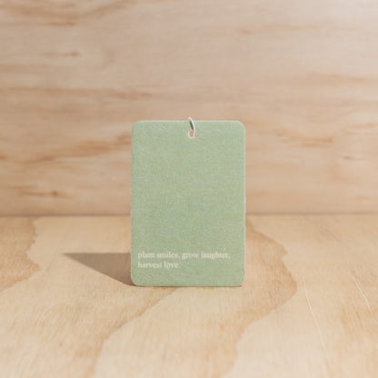 The Commonfolk air freshener features a green label with white text that reads 'plant smiles, grow laughter, harvest love' displayed on a wooden surface.