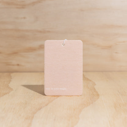 The Commonfolk air freshener features a pink label with a message that reads 'Stay in Your Magic' displayed on a wooden surface. 