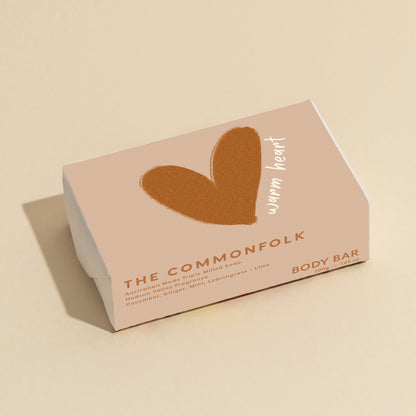 The Commonfolk 200g body bar soap, with a brown heart shape on the top, has packaging in a pinkish hue with the text 'warm heart.'